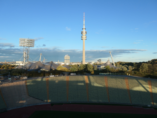 Das Olympiastadion in Berlin, Deutschland, mit dem Fernsehturm im Hintergrund, umgeben von Bäumen, Gebäuden und Lichtern, unter einem bewölkten Himmel.