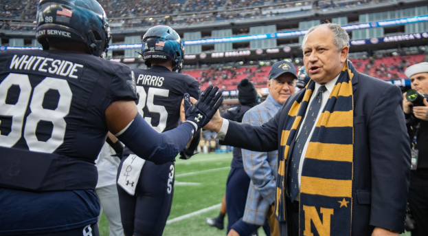 Ein Mann im Anzug und Krawatte gibt einem Fußballspieler die Hand auf einem Feld, umgeben von einer Gruppe von Menschen, mit einer Person, die eine Kamera auf der rechten Seite hält, und einem Stadion voller Menschen im Hintergrund, mit Schildern, auf denen "Notre Dame Football Coach Mike McCarthy und die Fighting Irish" steht.