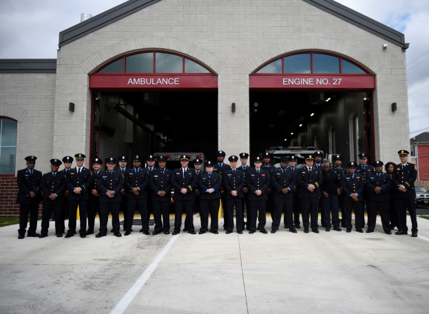 Eine Gruppe von Feuerwehrleuten in Uniform steht vor einer Feuerwache, mit Gras, Fahrzeugen, Gebäuden, Drähten und einem klaren blauen Himmel im Hintergrund.