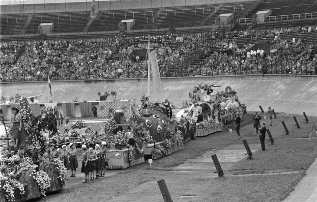 Schwarzes und weißes Foto einer Parade in einem Stadion mit Menschen, die stehen und sitzen, einem zentralen Springbrunnen und Blumensträußen auf den Fahrzeugen.