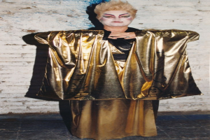 Eine Frau in einem goldenen Kimono posiert selbstbewusst vor einer weißen Backsteinwand, trägt ein schwarzes und goldenes Kleid mit einem glatten Bob-Haarschnitt, kräftigem Make-up und einem entschlossenen Gesichtsausdruck.
