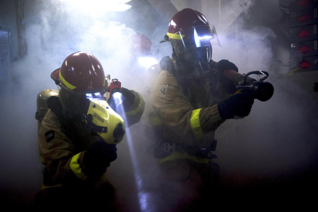 Zwei Feuerwehrleute in Schutzausrüstung spritzen Wasser auf eine rauchgefüllte Wand, mit einer Platte und verstreuten Gegenständen in der Nähe.