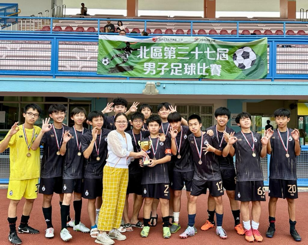 Gruppe junger Männer in Fußballtrikots auf einem Feld stehend, Medaillen tragend und einen Pokal haltend, mit einem 'Yokohama U-16 Jungenfußballteam' Banner im Hintergrund und Zuschauern in der Nähe sitzend.