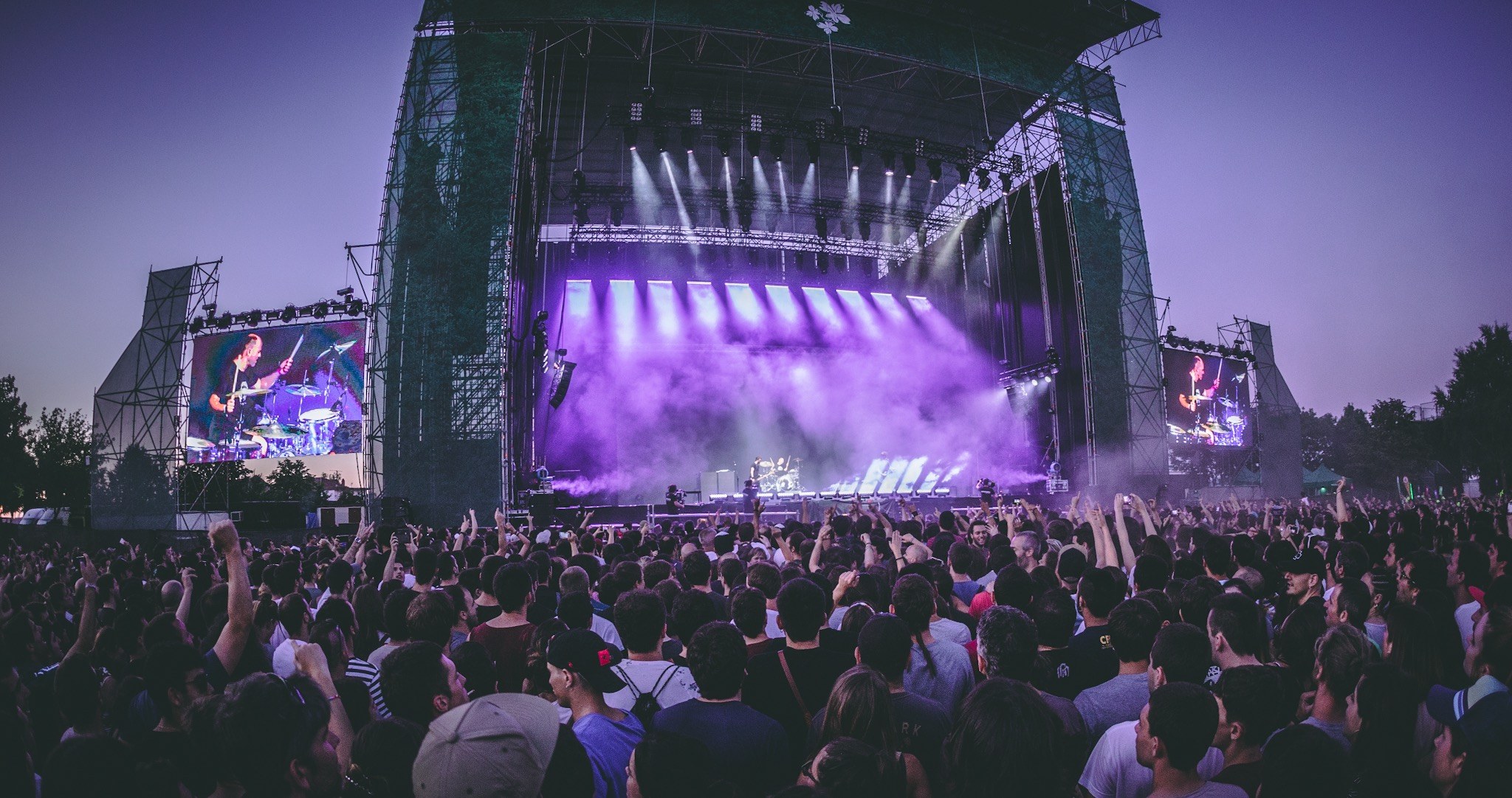 Eine große Menschenmenge auf einem Musikfestival schaut Künstlern auf einer hell erleuchteten Bühne mit Instrumenten und Bildschirmen zu, die vor einem Gebäude, Bäumen und einem klaren blauen Himmel steht.