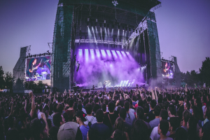 Eine große Menschenmenge auf einem Musikfestival schaut Künstlern auf einer hell erleuchteten Bühne mit Instrumenten und Bildschirmen zu, die vor einem Gebäude, Bäumen und einem klaren blauen Himmel steht.