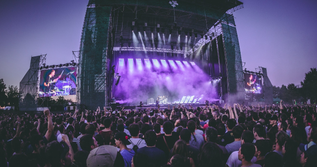 Eine große Menschenmenge auf einem Musikfestival schaut Künstlern auf einer hell erleuchteten Bühne mit Instrumenten und Bildschirmen zu, die vor einem Gebäude, Bäumen und einem klaren blauen Himmel steht.