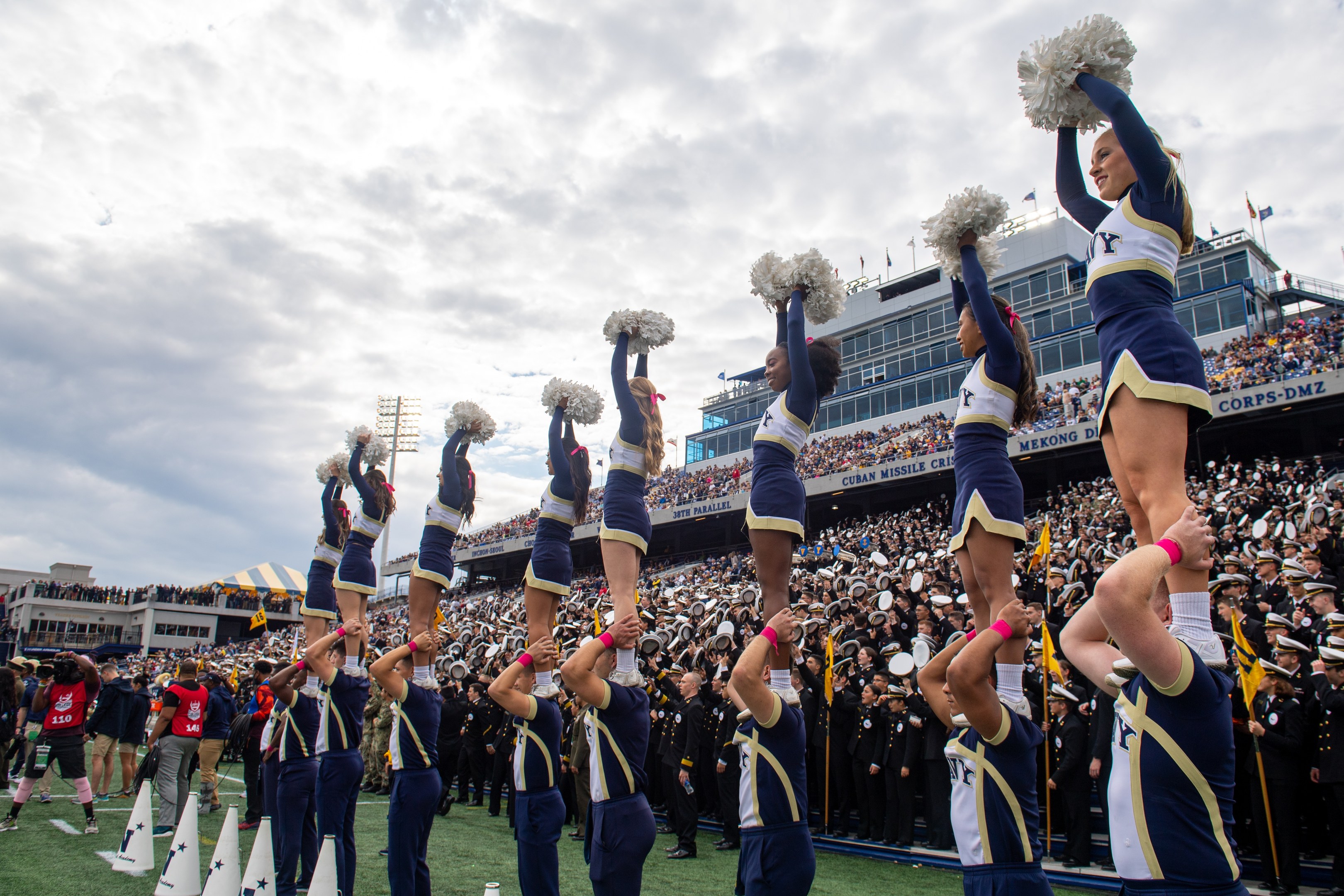Eine Gruppe von Cheerleadern in blauen und weißen Uniformen führt einen Stunt auf einem Stadionevent durch, während sie Pompons halten und eine Menge zusieht und eine Person die Szene fotografiert.
