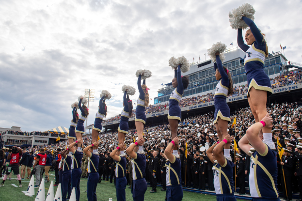 Eine Gruppe von Cheerleadern in blauen und weißen Uniformen führt einen Stunt auf einem Stadionevent durch, während sie Pompons halten und eine Menge zusieht und eine Person die Szene fotografiert.