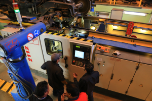 Vier Männer stehen um eine Maschine mit einem Bildschirm und Tasten in einer Fabrik, mit zusätzlichen Maschinen im Hintergrund.