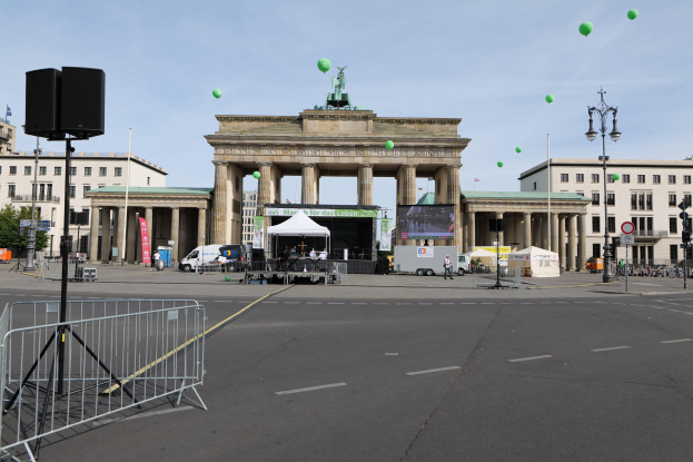 Das Brandenburgertor in Berlin, Deutschland, mit seinen ikonischen Säulen und Statuen, umgeben von Gebäuden, Laternenmasten, Verkehrssignalen, Zelten, Fahrzeugen, Menschen, Fahrrädern und grünen Ballons im bewölkten Himmel während des Berlin-Marathons.
