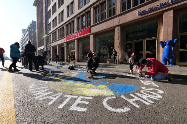 Menschen sitzen vor einem Gebäude mit Fenstern und Namensschildern auf dem Boden, umgeben von Flaschen und anderen Gegenständen, während sie an einer Klimawandel-Demonstration in Berlin teilnehmen, bei der es Bäume und einen klaren blauen Himmel gibt.