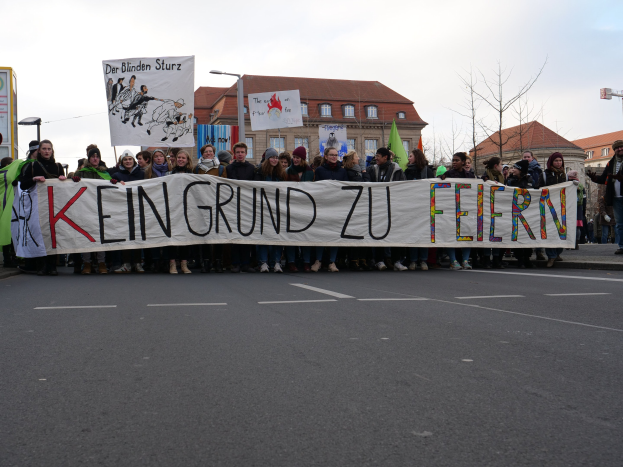 Gruppe von Menschen mit einem Banner mit der Aufschrift "Kein Grund zu Feiern" auf der Straße in Protest vor einem Gebäude mit Fenstern, Laternen, Bäumen und einem klaren blauen Himmel im Hintergrund.