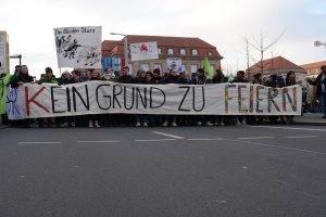 Demonstranten mit einem Banner "Kein Grund zu Feiern" gegen deutsche Sparmaßnahmen, auf einer Straße mit Gebäuden, Bäumen und einem klaren Himmel im Hintergrund.
