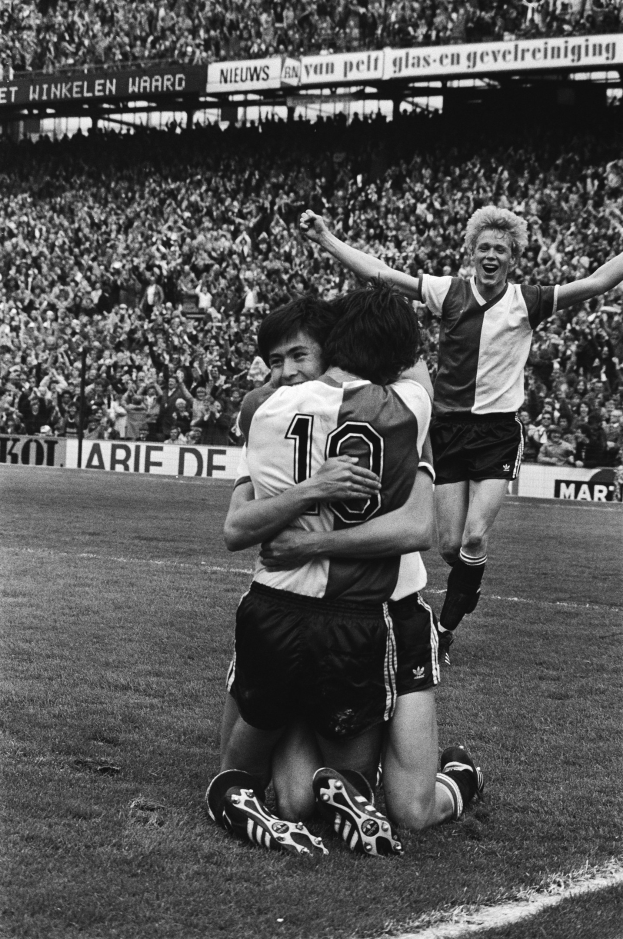 Drei Fußballer umarmen sich auf dem Boden nach einem Tor, mit jubelnder Menge und schwenkenden Fahnen im Hintergrund, in Schwarz-Weiß aufgenommen.