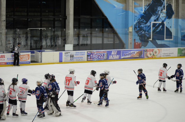 Gruppe junger Menschen in Helmen beim Eishockey auf einer Indoor-Eisfläche mit Bannern, Säulen und Glaswänden im Hintergrund.