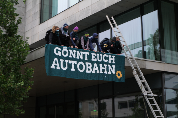 Menschen auf einem Gebäude stehend, die eine Fahne halten, mit einem Baum links und Glasfenstern im Hintergrund, die scheinbar für die deutsche Regierung protestieren.