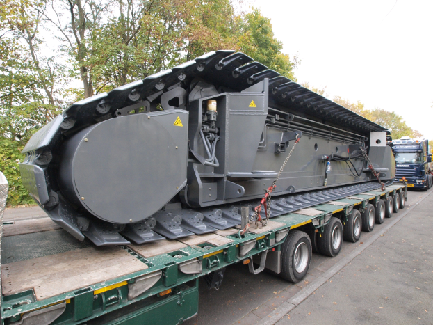Ein großer Lkw mit einer großen Maschine auf dem Rücken fährt eine von Bäumen gesäumte Straße unter einem klaren blauen Himmel entlang.