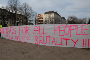 Eine Gruppe von Menschen hält ein Transparent mit der Aufschrift "Rechte für alle Menschen Stoppt Polizeigewalt" auf dem Boden mit einem Straßenschild, einem Schild, Bäumen, Gebäuden mit Fenstern und einem bewölkten Himmel im Hintergrund.