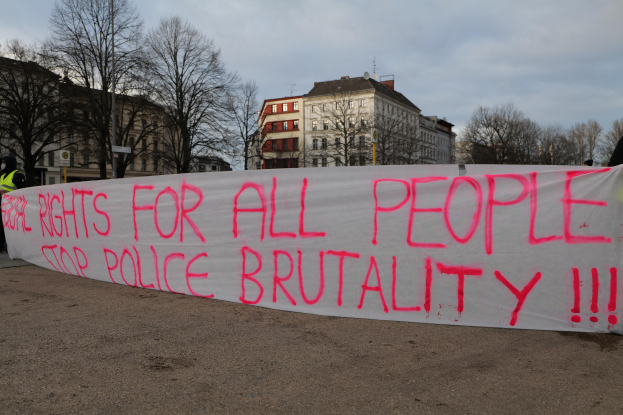 Eine Gruppe von Menschen hält ein Transparent mit der Aufschrift "Rechte für alle Menschen Stoppt Polizeigewalt" auf dem Boden mit einem Straßenschild, einem Schild, Bäumen, Gebäuden mit Fenstern und einem bewölkten Himmel im Hintergrund.
