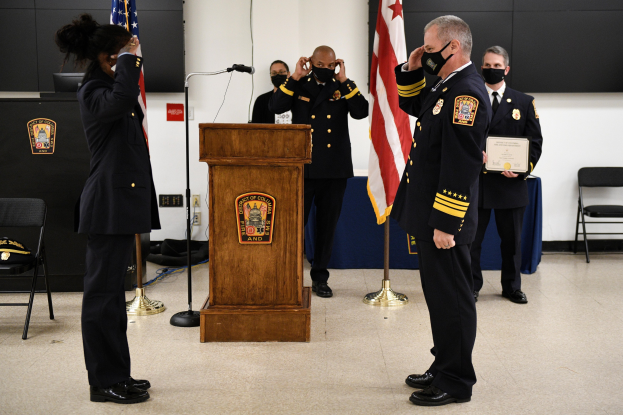 Eine Gruppe uniformierter und maskierter Personen steht vor einem Podium mit Mikrofon, umgeben von Fahnen, Stühlen und Tischen, während einer Vereidigung eines neuen Feuerwehrchefs, wobei eine Person ein Zertifikat hält.