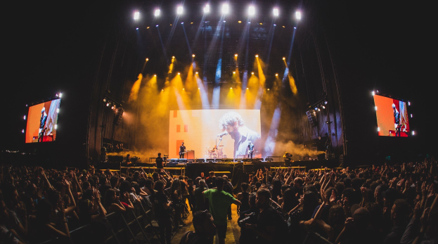 Eine große Menge Menschen steht vor einer hell erleuchteten Bühne, auf der Musiker Instrumente spielen und ein Sänger ein Mikrofon hält, umgeben von Bildschirmen und einer dunklen Kulisse.