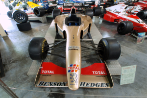 Rennwagen in einem Motorsportmuseum ausgestellt, durch einen Metallzaun von Zuschauern getrennt, mit informativen Texten im Hintergrund.