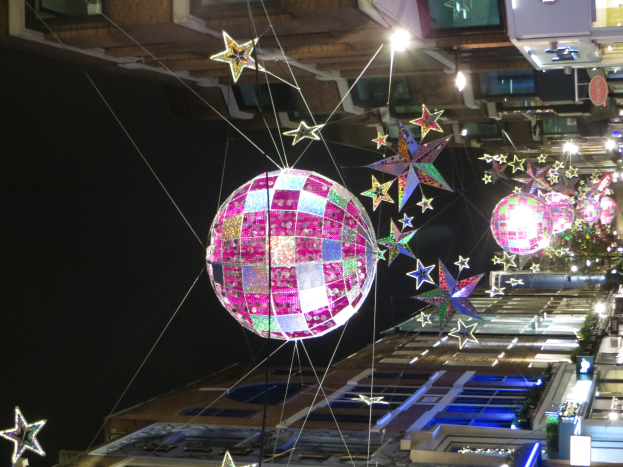 Eine Stadtstra├če bei Nacht mit zahlreichen Lichtern und Dekorationen, darunter eine gro├če Diskokugel, die von einem Geb├Ąude h├Ąngt und eine festliche Atmosphäre schafft.