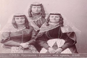 Drei Frauen in traditionellen russischen Hochzeitskleidern mit aufwendiger Stickerei und Perlen stehen vor einer weißen Wand in einem Schwarz-Weiß-Foto.